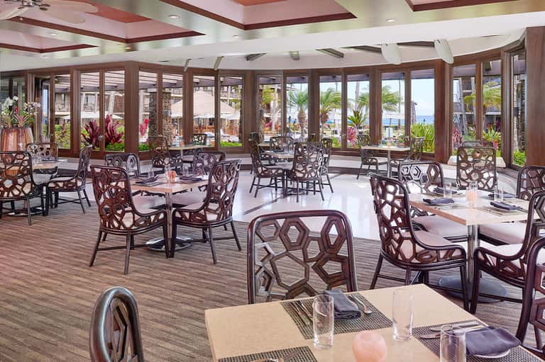 Upscale dining room with ocean view windows, elegant chairs, and tropical flowers overlooking beach
