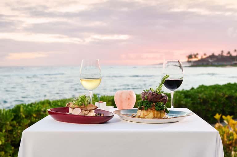 Plated dinner with wine glasses overlooking sunset ocean and tropical foliage