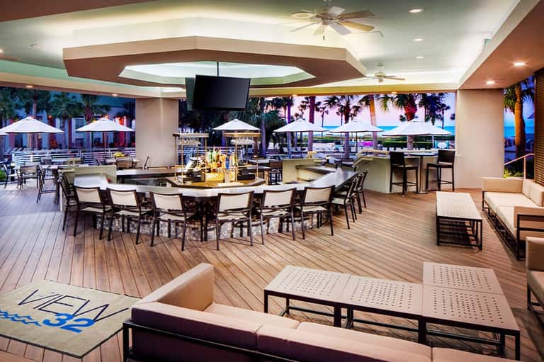 Seaside terrace lounge with bar, dining areas, white umbrellas, palm trees, and ocean views