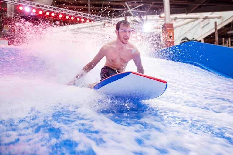 Man surfing on indoor wave machine with foam, blue surfboard, and red lighting overhead