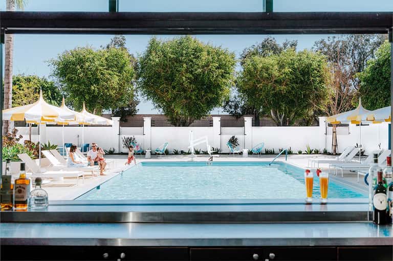 Indoor-outdoor pool view with lounge chairs, white umbrellas, trees, and swimmers visible from above
