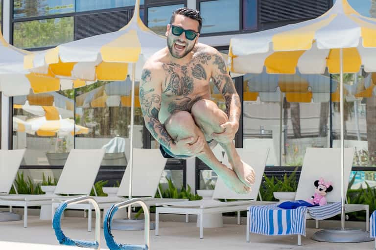 Tattooed man holding child by pool with yellow and white striped umbrellas in background