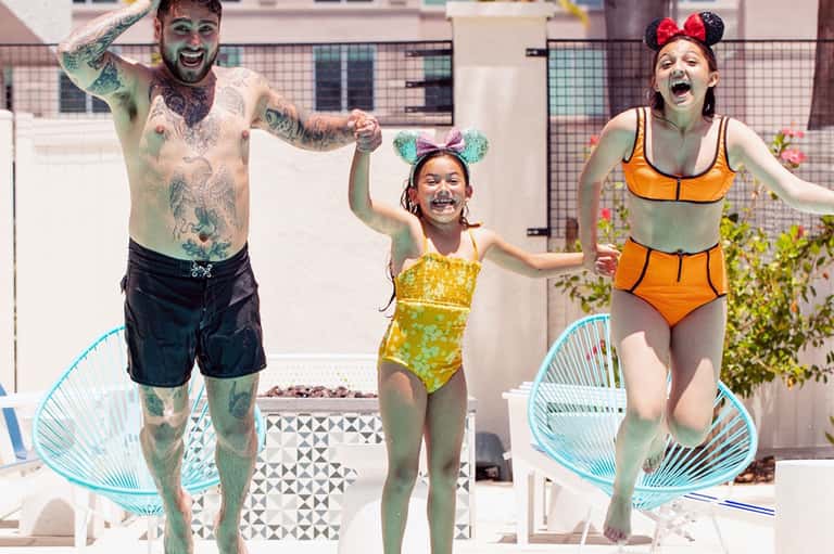 Family of three celebrating in pool wearing Mickey ears and colorful swimwear on rooftop deck