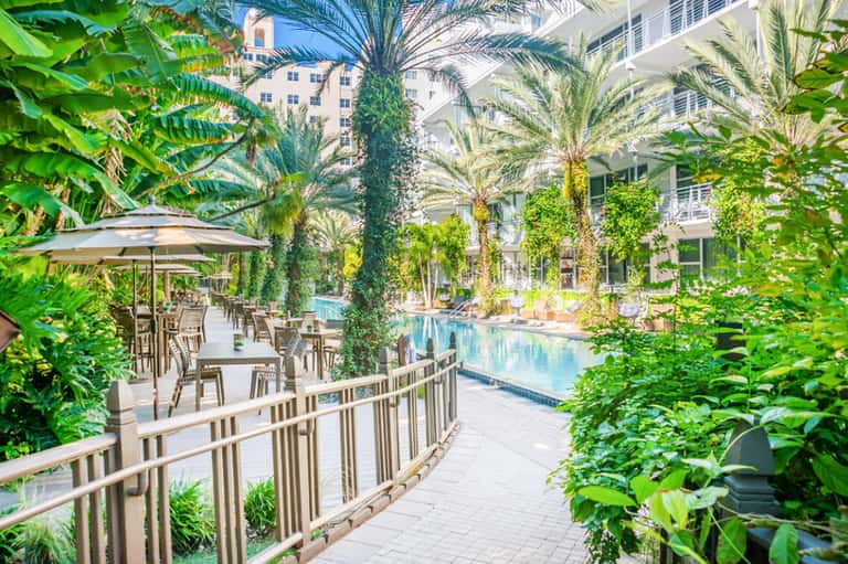 Lush tropical garden pathway with palm trees, greenery, umbrellas, and modern resort building entrance