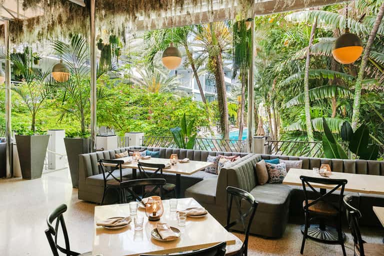 Indoor dining space with green tropical plants, natural lighting, and gray upholstered seating near tables