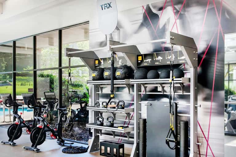 Modern fitness center with exercise bikes, weight equipment, TRX straps, and green outdoor view