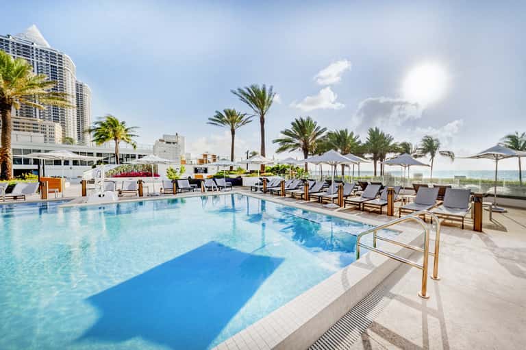 Rooftop outdoor pool with turquoise water, palm trees, lounge chairs, umbrellas, and high-rise building backdrop.