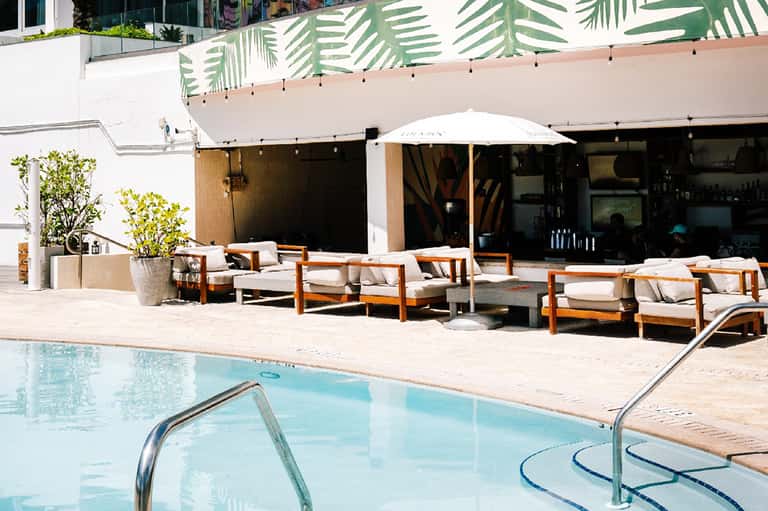 Modern poolside lounge with wooden furniture, white umbrella, and tropical foliage backdrop