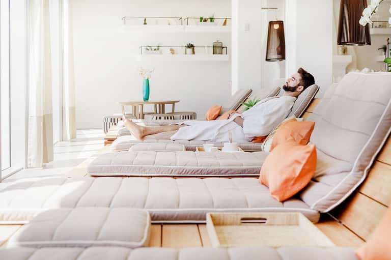 Man relaxing on white spa lounger with orange accent pillows in minimalist wellness room with natural light