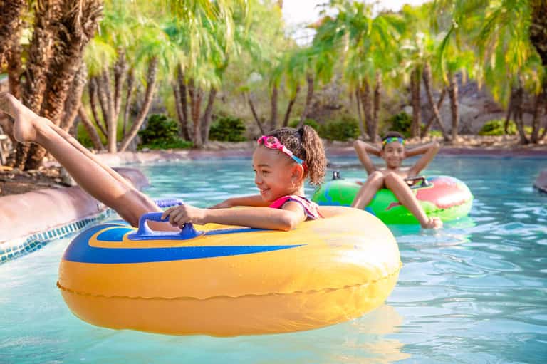 Children playing on colorful inflatable tubes in a tropical resort pool surrounded by palm trees