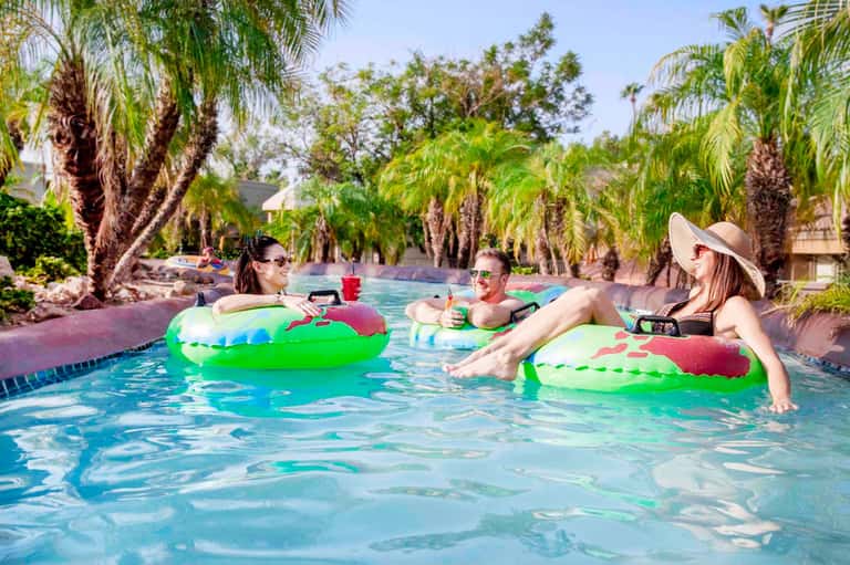 Four women relaxing on colorful inflatable floats in a tropical resort pool surrounded by palm trees