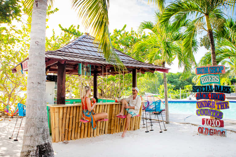 Beachside tiki bar with wooden shelter, colorful signage, and guests relaxing by the pool with palm trees