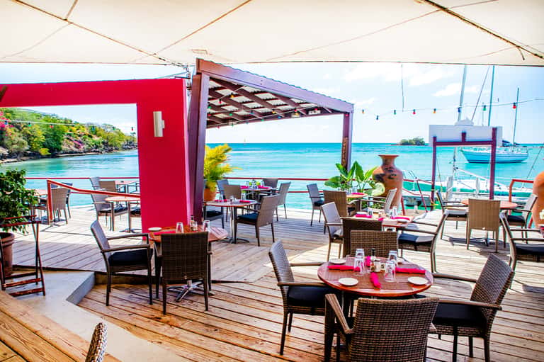 Beachfront dining area with red accents, wicker chairs, wooden deck, and sailboats visible in turquoise water