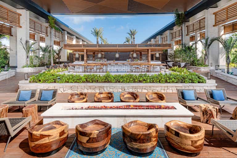 Modern courtyard with fire pit, wooden seating, bar area, and palm trees at midday