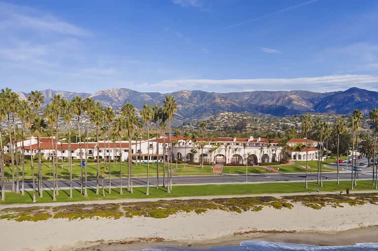 Beachfront property with Spanish-style white building, palm trees, and mountains in background