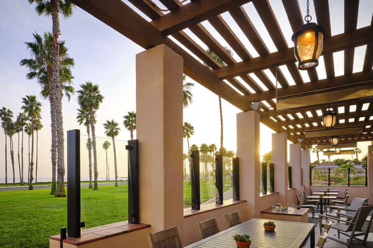 Covered outdoor dining area with pergola, palm trees, and manicured lawn at dusk