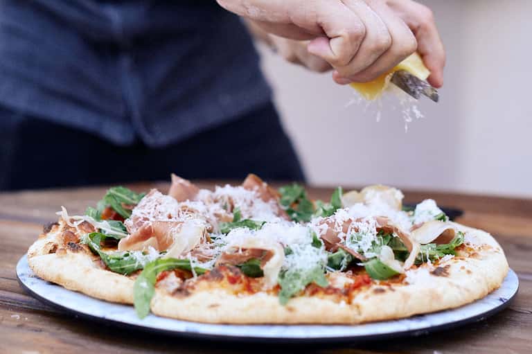 Chef grating parmesan cheese over fresh pizza with prosciutto and greens