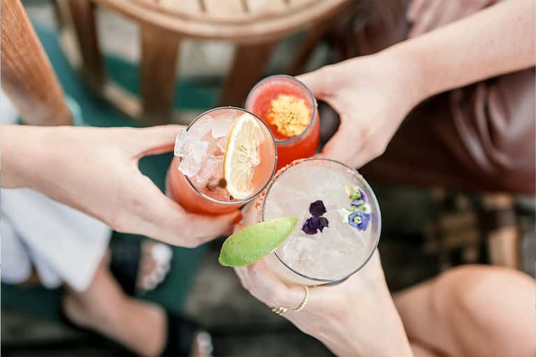 Hands holding three colorful cocktails garnished with citrus and edible flowers