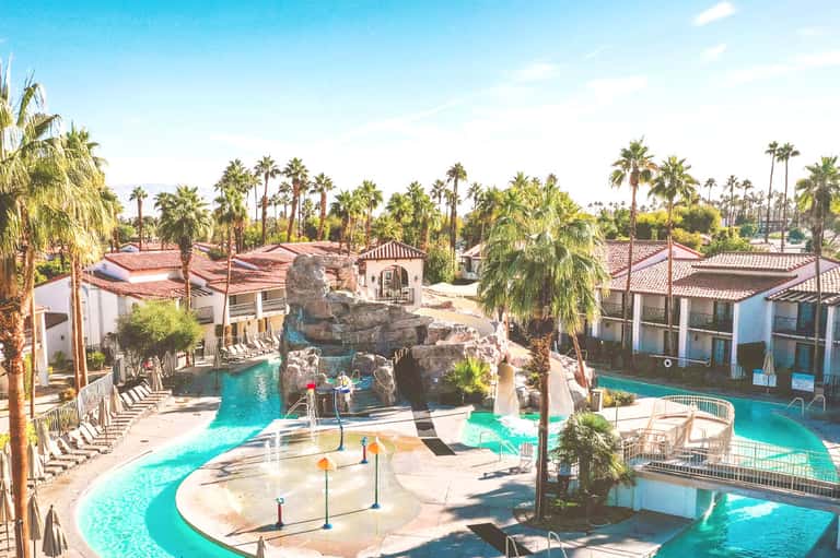 Tropical resort pool area with rock formations, palm trees, turquoise water, and Spanish architecture