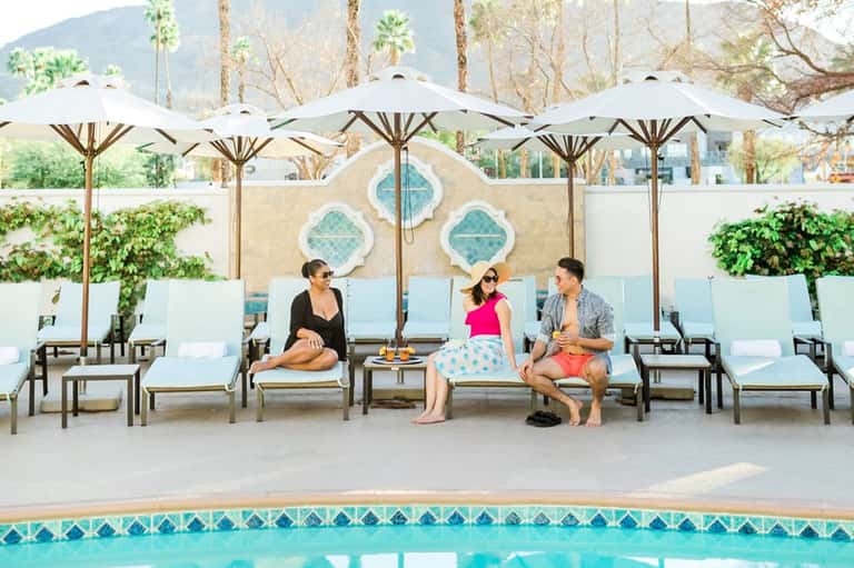 Three guests lounging by pool under white umbrellas with decorative cushions and mountains