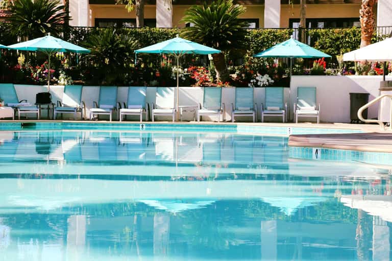 Resort pool with turquoise umbrellas, white lounge chairs, flowering hedges, and palm trees
