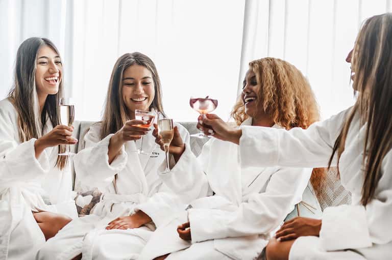 Four women in white spa robes toasting with champagne glasses in bright, modern spa setting