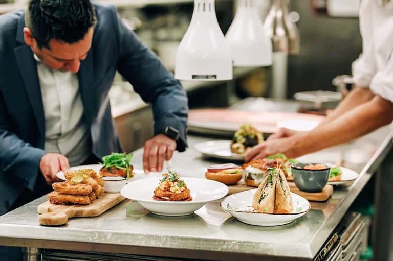 Chef plating gourmet dishes in professional kitchen with white pendant lights and stainless steel counters
