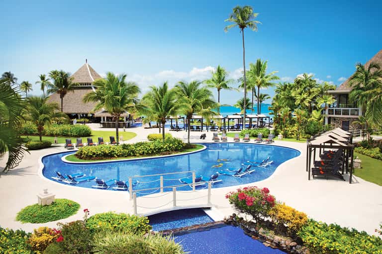 Curved resort pool with thatched roof pavilion, palm trees, and ocean view in tropical setting