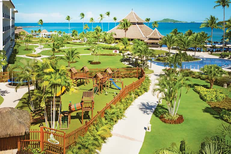 Tropical resort overview with green lawns, thatched structures, pools, and ocean horizon