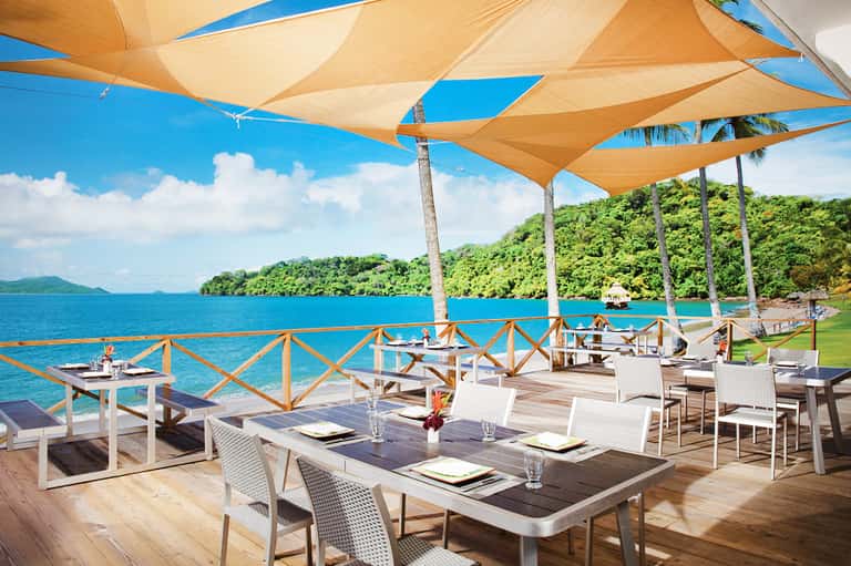 Beachfront dining area with wooden railings, shade sails, and turquoise water views