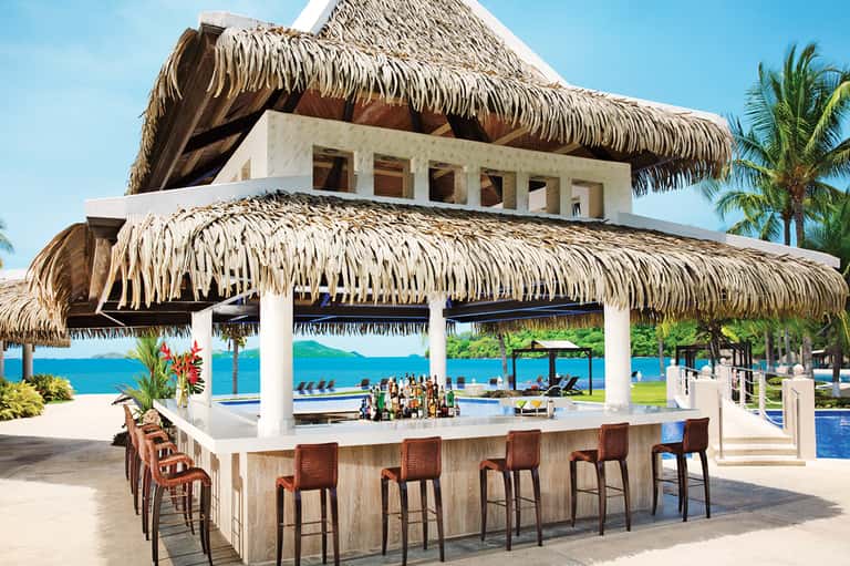 Beachfront bar with thatched roof, white columns, pool view, and blue ocean horizon