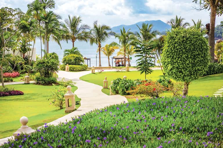 Manicured tropical gardens with flowering plants, palm trees, and mountain water views