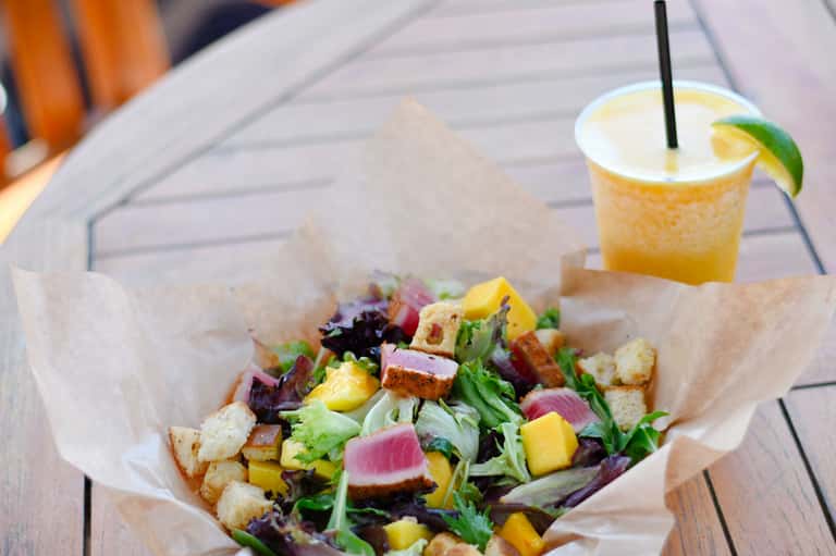 Colorful salad with seared tuna, greens, and croutons in paper wrapper with citrus cocktail on wooden deck