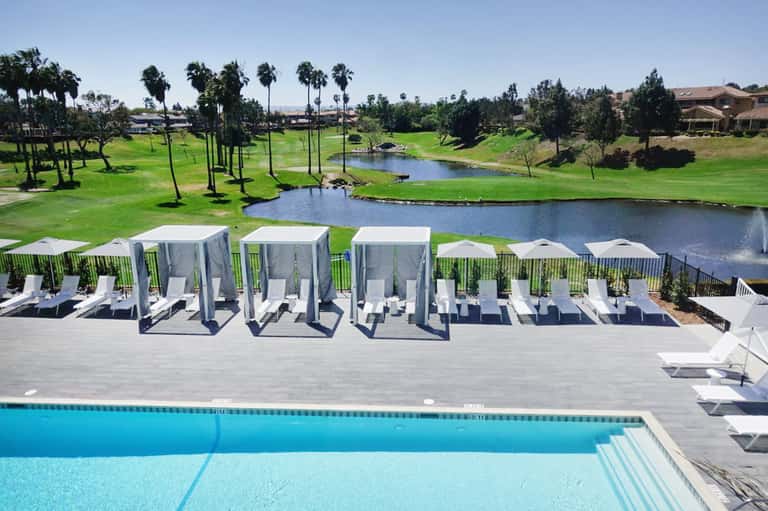 Resort pool deck with white cabanas overlooking a golf course and lake under clear blue sky