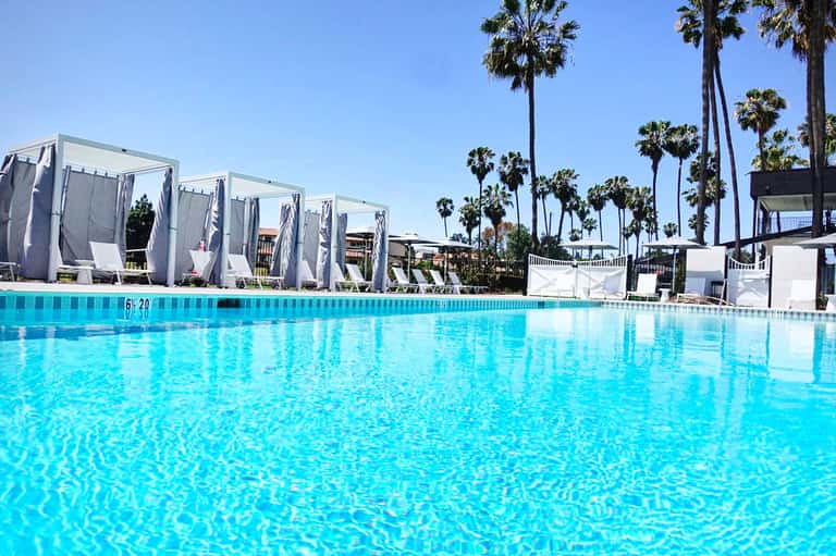 Bright turquoise resort pool with white cabanas and tall palm trees under clear blue sky