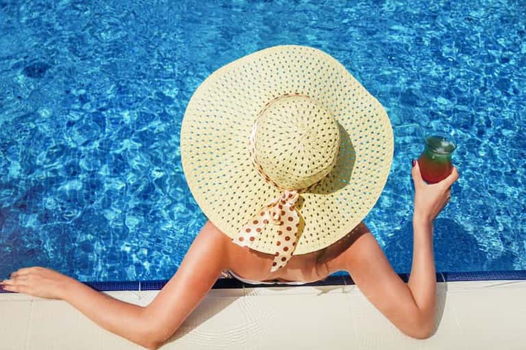 Woman in wide-brimmed straw hat lounging at pool edge holding colorful cocktail