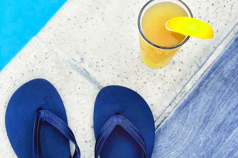 Yellow cocktail with lime garnish by poolside, blue flip-flops on concrete deck