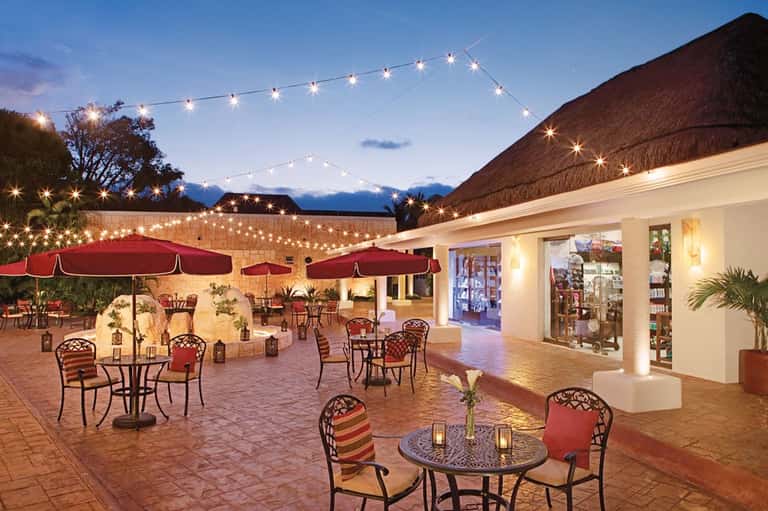 Outdoor courtyard at dusk with red umbrellas, thatched roofs, string lights, and mountain views