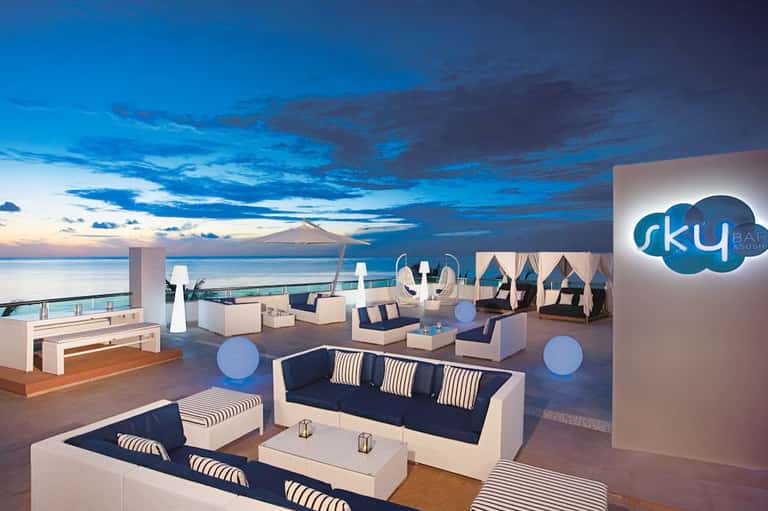 Luxury rooftop bar at dusk with ocean views, modern furnishings, and blue accent lighting overlooking the coastline