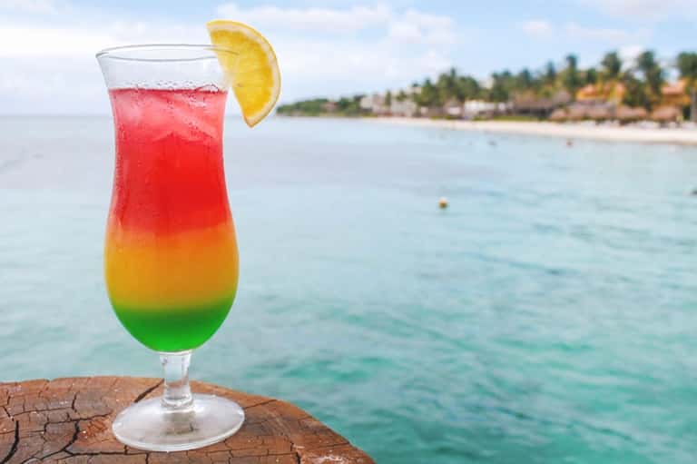 Rainbow layered cocktail with lemon slice on a wooden dock overlooking tropical beach