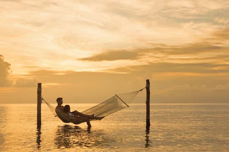 Couple relaxing in hammock at sunset over calm ocean water during golden hour