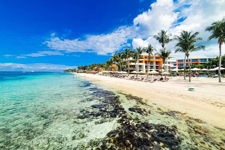 Tropical beach resort with turquoise water, sandy beach, colorful buildings, palm trees, and lounge chairs