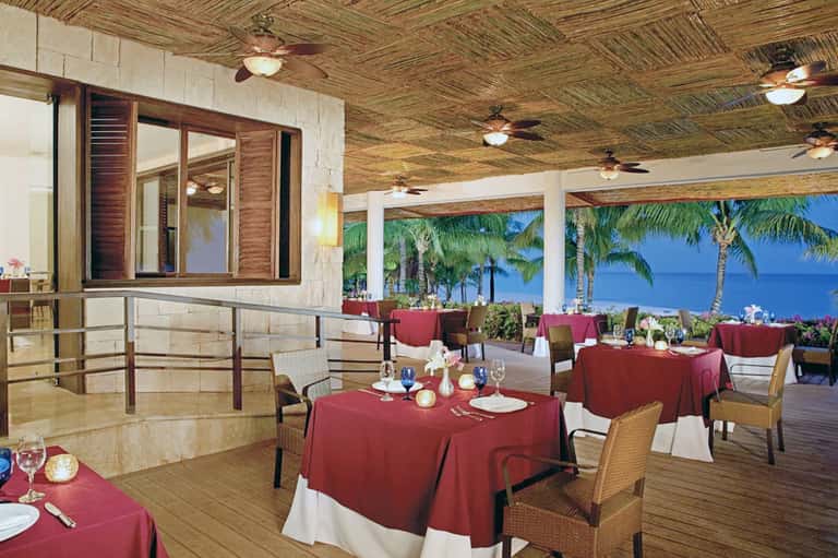 Upscale beachfront dining room with red tablecloths, wicker chairs, ocean views, and palm trees