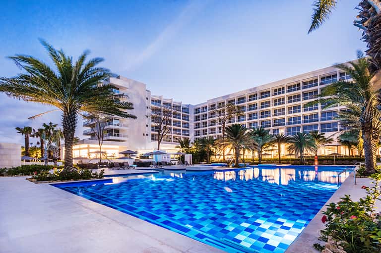 Modern resort at dusk with illuminated pool, palm trees, and multi-story building