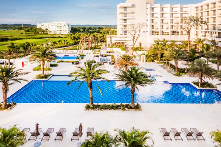 Aerial view of resort with multiple blue pools, white buildings, palm trees, and green surroundings