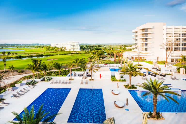 Aerial view of resort with multiple blue pools, palm trees, and lush green marshland surroundings