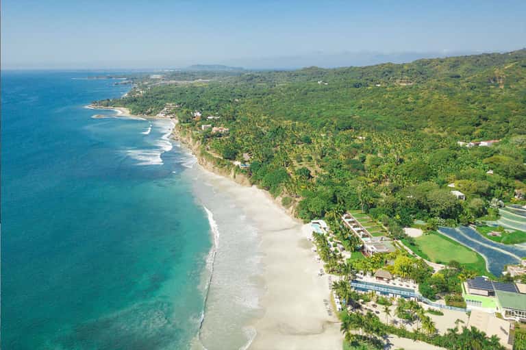 Aerial view of pristine beach with turquoise waters, lush green hillside, and luxury coastal development