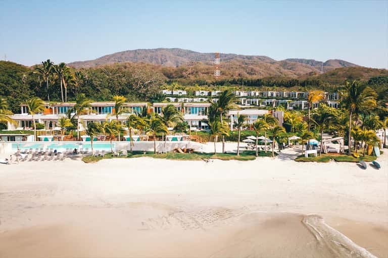 Beachfront resort with white buildings, pools, and palm trees backed by mountains