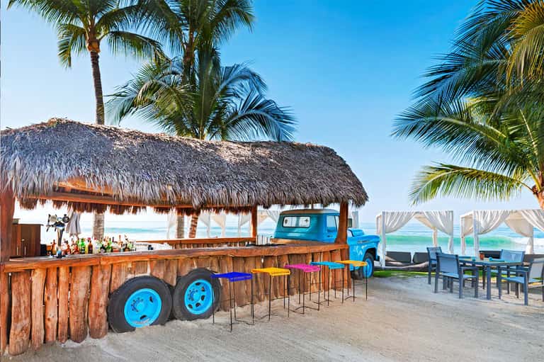 Beachfront tiki bar with thatch roof, blue truck decor, colorful stools, and ocean view