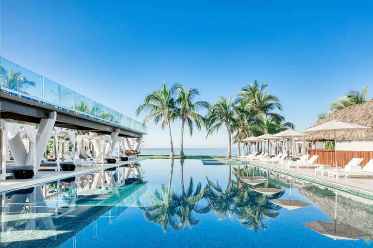 Infinity pool reflecting palm trees and ocean with white cabanas and covered lounging areas
